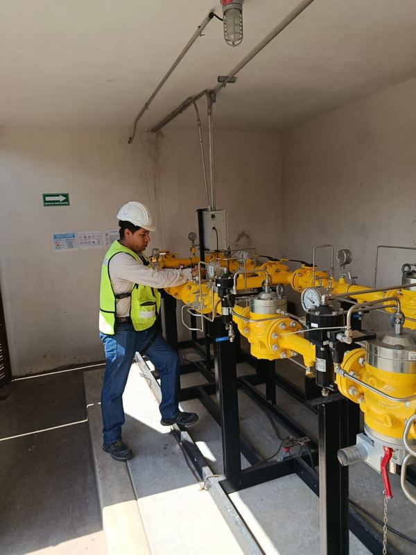 Técnico con casco y chaleco de alta visibilidad operando válvulas de un tren de regulación y medición amarillo dentro de una caseta