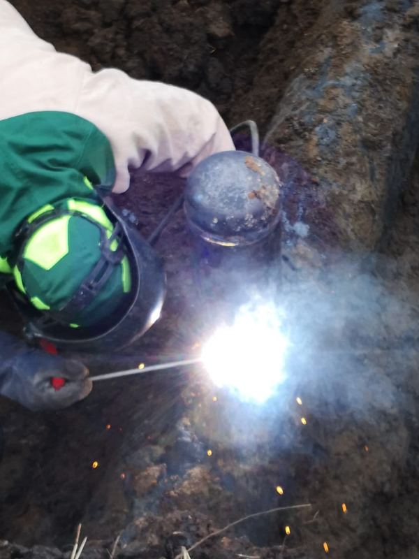 Soldador con careta aplicando soldadura de arco sobre una conexión de tubería de acero dentro de una zanja