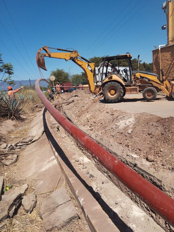 Retroexcavadora junto a una zanja mientras se tiende una tubería de acero con recubrimiento rojo