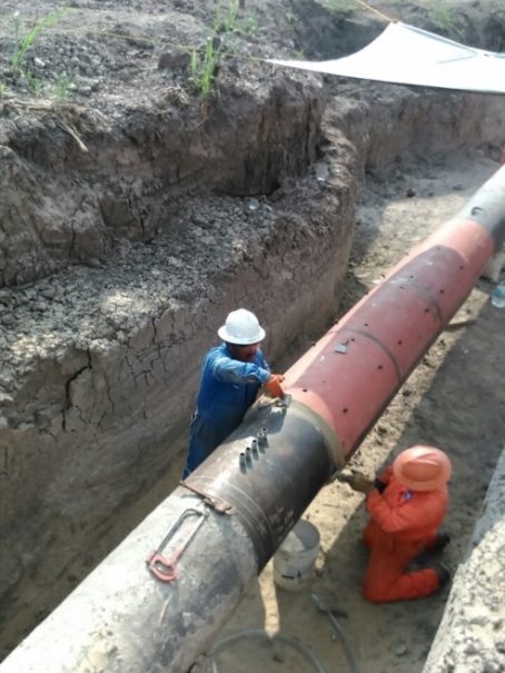 Dos trabajadores aplicando recubrimiento a la junta de una tubería de acero dentro de una zanja
