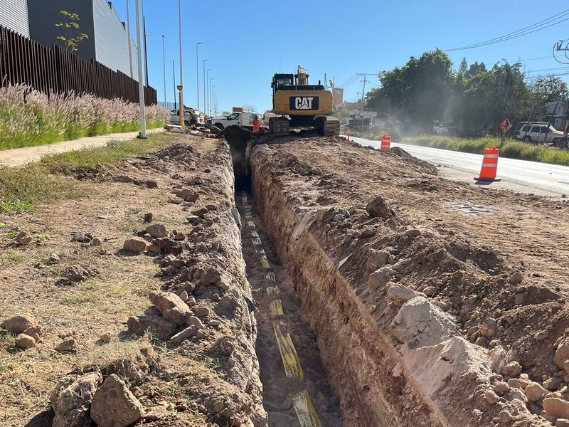 Zanja recta junto a una vialidad con cinta de advertencia de tubería de gas natural y una excavadora al fondo