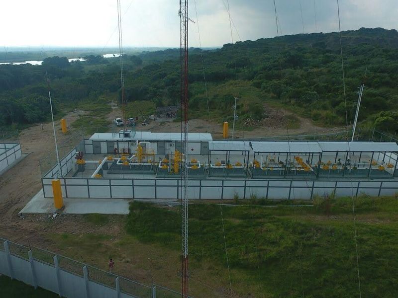 Vista aérea de una estación cercada con tuberías y equipos bajo techumbre, con vegetación al fondo