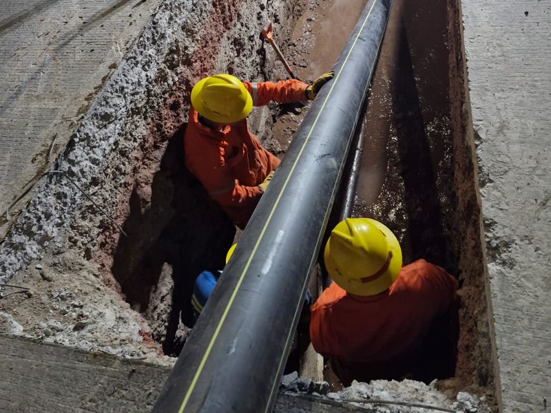 Dos trabajadores con casco amarillo y overol naranja dentro de una zanja abierta en pavimento colocando tubería de polietileno negra, de noche