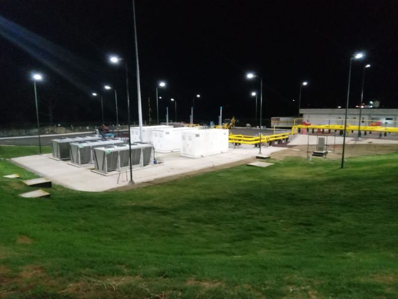 Instalación iluminada de noche con gabinetes de equipo, enfriadores y tubería amarilla, rodeada de áreas verdes